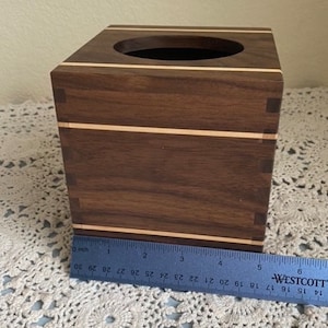 May include: A square wooden tissue box with a dark brown finish and light wood accents. The box has a circular opening on top and measures approximately 6 inches in length. The box is sitting on a white lace surface.