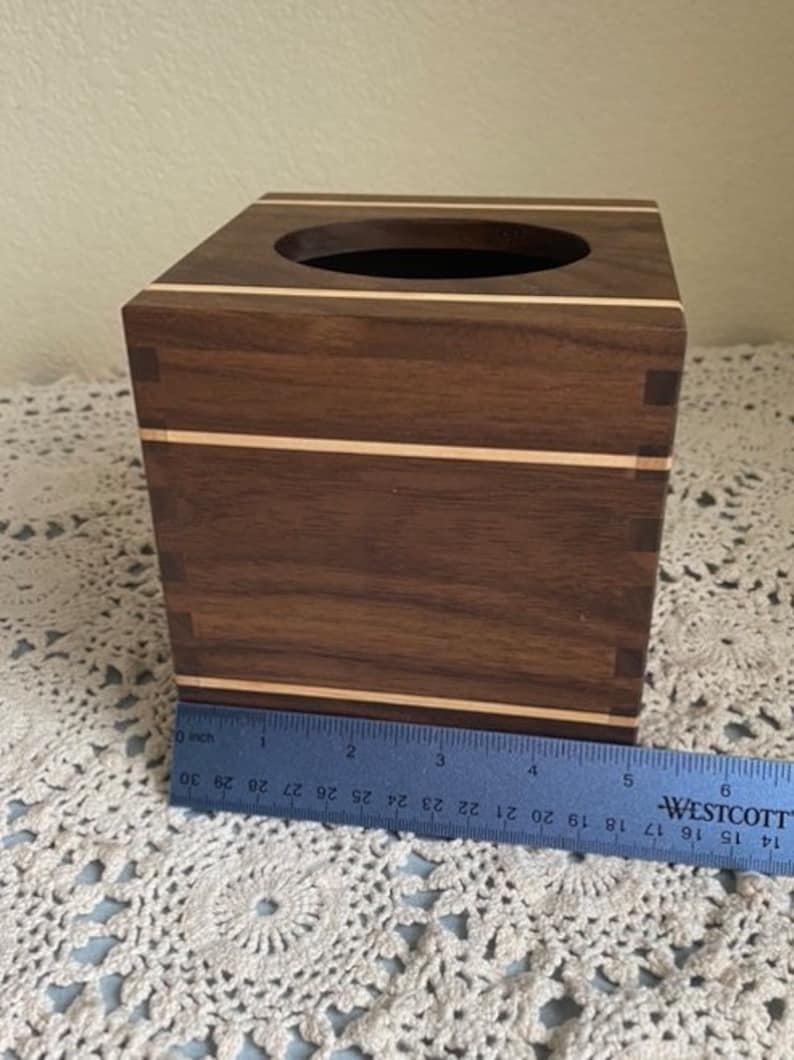 May include: A square wooden tissue box with a dark brown finish and light wood accents. The box has a circular opening on top and measures approximately 6 inches in length. The box is sitting on a white lace surface.