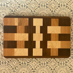 Puede incluir: Una tabla de cortar rectangular de madera con un patrón de tablero de ajedrez de madera clara y oscura. La tabla está hecha de madera de arce y nogal.
