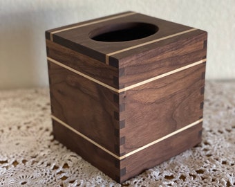May include: A square wooden tissue box with a dark brown finish and light-colored accent stripes. The box has a rectangular opening on top for dispensing tissues. The corners are joined with visible dovetail joints, showcasing the craftsmanship.
