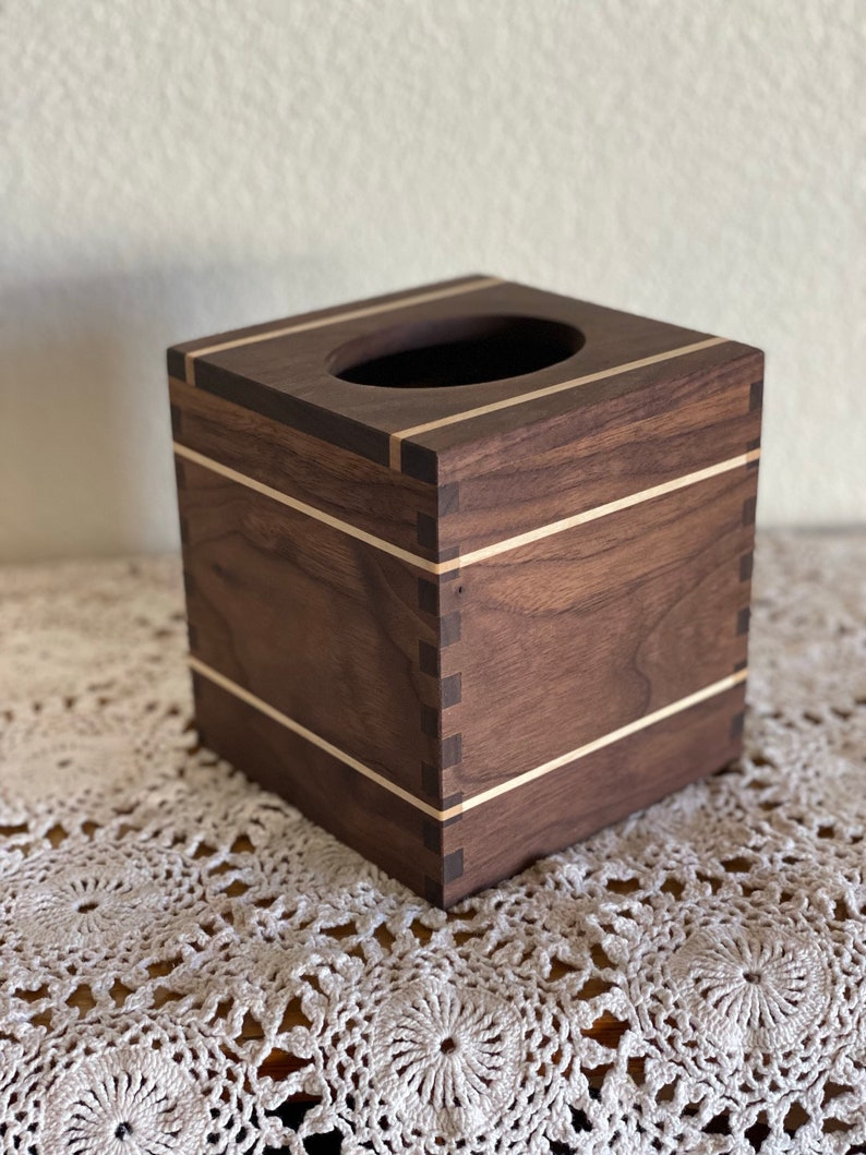 May include: A square wooden tissue box with a dark brown finish and light-colored accent stripes. The box has a rectangular opening on top for dispensing tissues. The corners are joined with visible dovetail joints, showcasing the craftsmanship.
