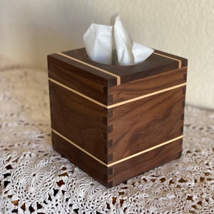 May include: A square wooden tissue box with a dark brown finish and light wood accents. The box has a simple, modern design with visible dovetail joints. White tissues are visible at the top.