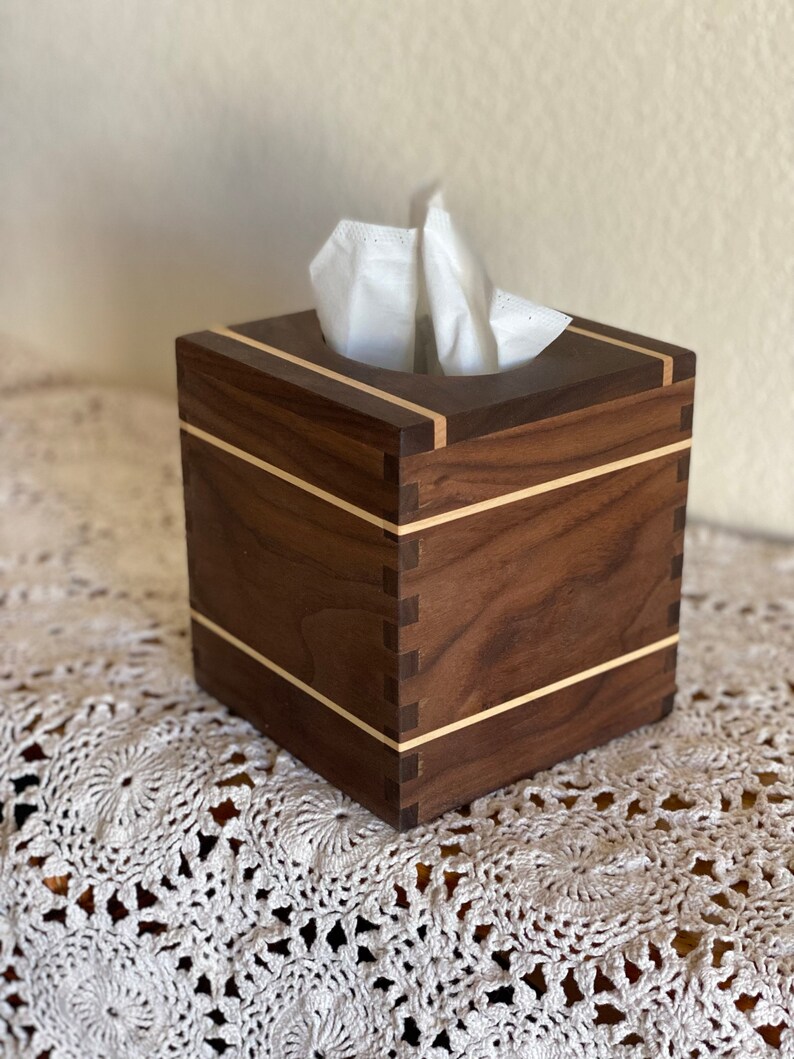 May include: A square wooden tissue box with a dark brown finish and light wood accents. The box has a simple, modern design with visible dovetail joints. White tissues are visible at the top.