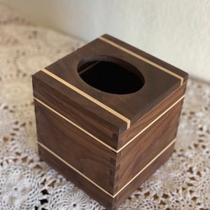 May include: A square wooden tissue box with a dark brown finish and light wood accents. The box has a rectangular opening on top and is constructed with visible dovetail joints. The light wood trim borders the top and bottom edges.