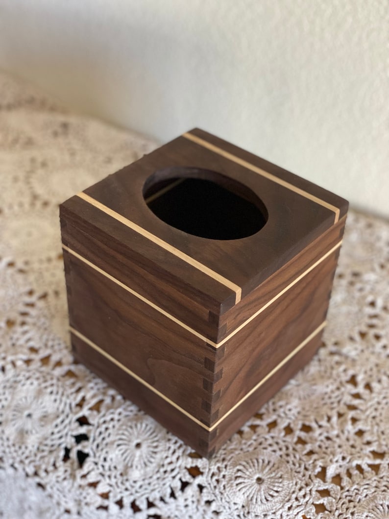 May include: A square wooden tissue box with a dark brown finish and light wood accents. The box has a rectangular opening on top and is constructed with visible dovetail joints. The light wood trim borders the top and bottom edges.