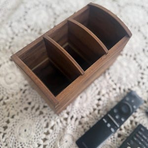 Può includere: Un organizer per telecomandi in legno con tre scomparti. L'organizer è di colore marrone caldo e ha una parte superiore curva. Due telecomandi neri sono visibili su una superficie di pizzo bianco.