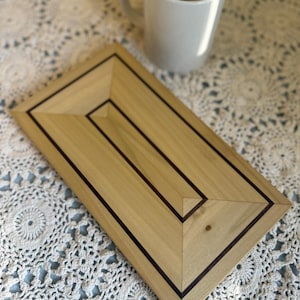 May include: A rectangular wooden tray with a light wood grain and dark brown inlay. A white mug sits to the side on a white lace tablecloth. The tray has a geometric design and is likely used for serving or display.
