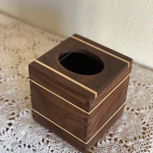 May include: A square wooden tissue box with a dark brown finish and light wood accents. The box has a rectangular opening on top and is constructed with visible dovetail joints. The design is simple and elegant.