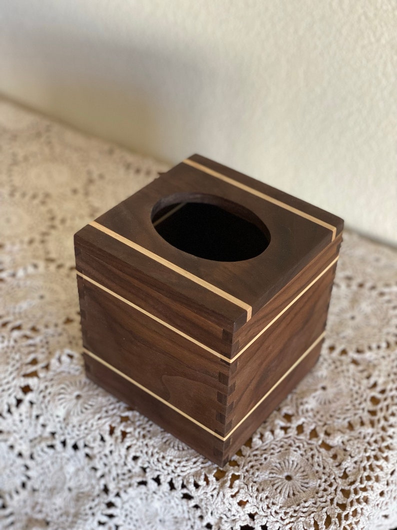 May include: A square wooden tissue box with a dark brown finish and light wood accents. The box has a rectangular opening on top and is constructed with visible dovetail joints. The design is simple and elegant.
