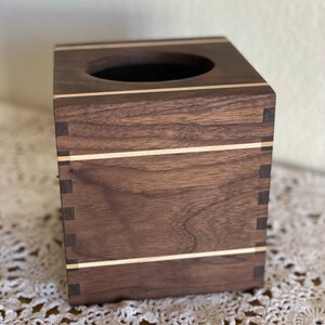 May include: A square wooden tissue box with a dark brown finish and light wood inlays. The box has a circular opening on top and is constructed with visible dovetail joints at the corners. The design is simple and elegant.