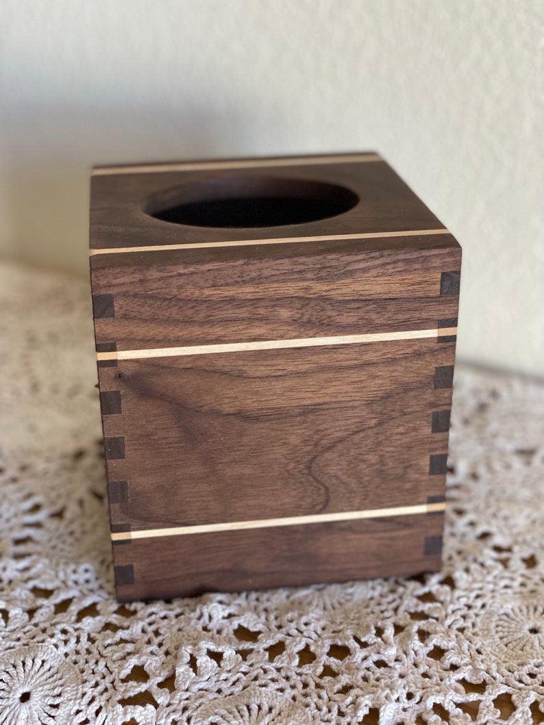 May include: A square wooden tissue box with a dark brown finish and light wood inlays. The box has a circular opening on top and is constructed with visible dovetail joints at the corners. The design is simple and elegant.