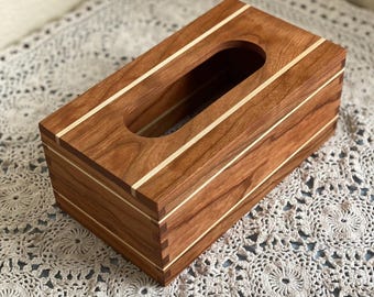 May include: A rectangular wooden tissue box with a rich brown finish and light-colored accent stripes. The box features a rectangular opening on top for easy tissue access. The design showcases a blend of natural wood tones and clean lines.