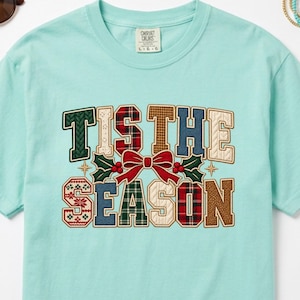May include: A light mint green short-sleeve t-shirt featuring the text TIS THE SEASON in large, multi-patterned block letters. The design includes a central red bow with green holly leaves and red berries, surrounded by various plaid and knit textures.