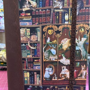 Puede incluir: Un diario cubierto de tela con un estampado de gatos entre estanterías. La cubierta es de un marrón intenso con un lomo a juego y un cierre elástico brillante. Las páginas interiores no son visibles.