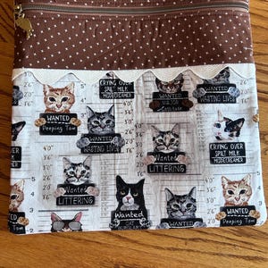 May include: A brown fabric zippered pouch with a white fabric lining. The lining features a repeating pattern of cats in a mugshot style with wanted posters. The text on the posters includes "Wanted", "Peeping Tom", "Wasting Lives", "Crying Over Spilt Milk", "Major Cattitude", "Littering", and "Cat Burglar".