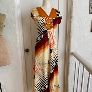 Puede incluir: Un vestido largo de maxi vintage con un patrón geométrico en tonos de marrón, naranja, blanco y negro. El vestido tiene un escote en V y mangas cortas. El corpiño presenta un material marrón similar al ante con un detalle de encaje.