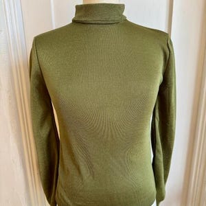 Puede incluir: Jersey de cuello alto verde oliva con mangas largas. El jersey se muestra en un maniquí. La tela tiene una sutil textura acanalada. El cuello alto está doblado.