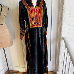 May include: A black velvet dress with long sleeves and a V-neck. The dress features intricate embroidery in red, yellow, and orange around the neckline and cuffs. Vertical orange stripes run down the front of the dress.