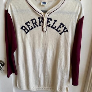May include: Cream Henley shirt with burgundy sleeves and trim. The word "BERKELEY" is arched across the chest in navy blue letters. The shirt has a three-button placket and a classic baseball style.