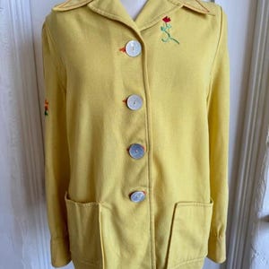 May include: A yellow jacket with a notched collar, two front pockets, and large, pearlescent buttons. The jacket features red stitching and embroidered floral accents. The jacket is displayed on a mannequin.