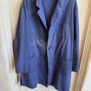 May include: A blue work coat with a button closure, two front pockets, and a chest pocket. The coat is hanging on a hanger against a white door. The coat has a collar and long sleeves.