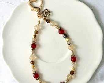 May include: A beaded keychain featuring a gold-toned heart-shaped lobster clasp and a small four-leaf clover charm. The loop consists of alternating round beads in shades of red, translucent beige, and brown, accented with small gold-toned star charms.