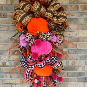 May include: A festive fall door swag with orange, pink, and black velvet pumpkins, leopard print ribbon, and black and white checkered ribbon.