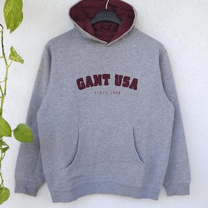 Può includere: Una felpa con cappuccio a maniche lunghe grigio melange con fodera interna bordeaux. La parte anteriore presenta la scritta GANT USA in lettere bordeaux, con il testo più piccolo SINCE 1949 sottostante. Include una tasca a marsupio frontale.