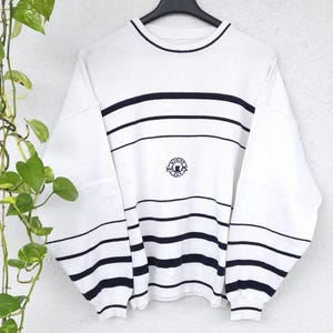 Peut inclure: Pull-over blanc à rayures horizontales bleu marine et col rond. Le pull-over porte un petit écusson brodé sur la poitrine et est suspendu à un cintre noir. Les manches sont longues et le pull est en matière douce.