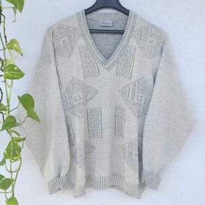 Peut inclure: Un pull col en V gris clair avec des motifs géométriques. Le pull est suspendu à un cintre noir contre un mur blanc. Il a des manches longues et des poignets et un ourlet côtelés. Une plante verte à feuilles à motifs est dans le coin supérieur gauche.