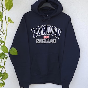 Könnte beinhalten: Ein dunkelblauer Kapuzenpullover mit dem Schriftzug LONDON ENGLAND in weißen Blockbuchstaben und einer kleinen Union-Jack-Flagge zwischen den Wörtern. Der Hoodie verfügt über eine Kängurutasche vorne und passende dunkelblaue Kordelzüge.