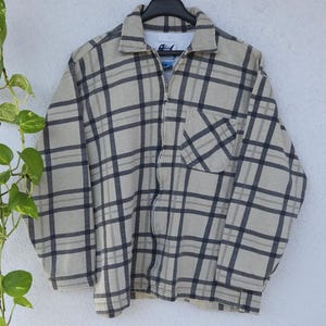 Könnte beinhalten: Beige-marine karierte Reißverschlussjacke, die an einem schwarzen Kleiderbügel hängt. Die Jacke hat einen Kragen, eine Vordertasche und einen durchgehenden Reißverschluss. Das Karomuster ist ein klassisches Design.