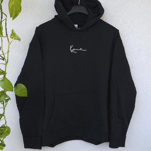 Könnte beinhalten: Ein schwarzer Kapuzenpullover mit langen Ärmeln und einem weißen, gestickten Signatur-Logo auf der Brustmitte. Das Kleidungsstück verfügt über eine Kängurutasche auf der Vorderseite, eine Kapuze mit Kordelzug sowie gerippte Ärmelbündchen.
