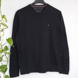 Könnte beinhalten: Ein schwarzer Rundhals-Pullover mit langen Ärmeln, hängt an einem schwarzen Kleiderbügel. Der Pullover hat ein kleines Logo auf der Brust und ein gestreiftes Detail am Halsausschnitt. Der Hintergrund ist eine schlichte weiße Wand.