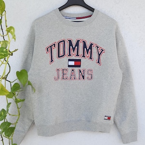 Könnte beinhalten: Ein meliertes graues Sweatshirt mit dem Schriftzug "TOMMY JEANS" in Marineblau und Rot. Das Sweatshirt hat ein kleines Logo unten rechts und hängt an einem schwarzen Kleiderbügel.