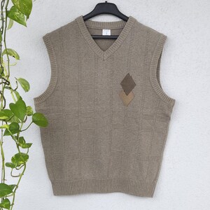 Peut inclure: Un gilet sans manches beige à col en V avec un motif en losanges et deux écussons en forme de losange marron. Le gilet est suspendu à un cintre noir contre un mur blanc, avec une plante verte en arrière-plan.