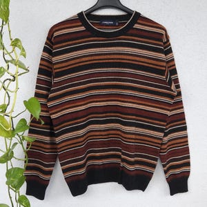 Peut inclure: Un pull à rayures marron, noir et beige est suspendu à un cintre noir. Le pull présente des rayures horizontales et des manches longues. Le mot "COMMANDER" est visible sur le col. Le pull est fait d'une matière douce.