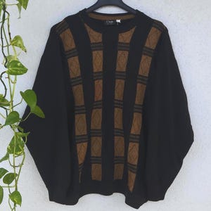 Könnte beinhalten: Ein schwarzer Pullover mit einem braunen geometrischen Muster. Der Pullover hängt an einem schwarzen Kleiderbügel vor einer weißen Wand. Das Muster besteht aus vertikalen braunen Streifen mit Rauten.