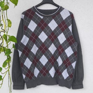 Peut inclure: Un pull gris à motif losanges avec des diamants blancs et bordeaux. Le pull a des manches longues et un col rond avec une bordure blanche. Le pull est suspendu à un cintre noir.