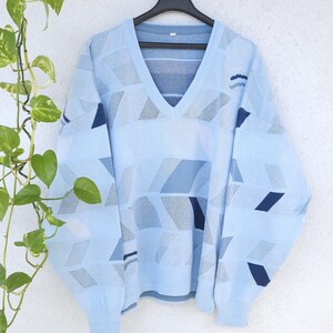 Könnte beinhalten: Hellblauer V-Ausschnitt-Pullover mit geometrischem Muster in Blau- und Grautönen. Der Pullover hängt an einem schwarzen Kleiderbügel vor einer weißen Wand, mit einer grünen Pflanze im Hintergrund. Das Design zeigt Rauten und Rechtecke.