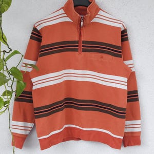 Könnte beinhalten: Orange-braun gestreifter Pullover mit einem Kragen mit Viertelreißverschluss. Der Pullover hat horizontale Streifen in Orange, Weiß und Braun. Er hat eine kleine Tasche auf der Brust und hängt an einem Kleiderbügel.