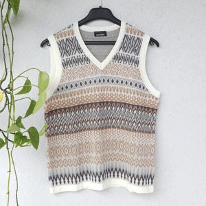 May include: A cream-colored, V-neck sweater vest with a geometric pattern in shades of brown, gray, and cream. The vest is sleeveless and hangs on a black hanger against a white wall. The brand name "GOLDNER" is visible on the label.