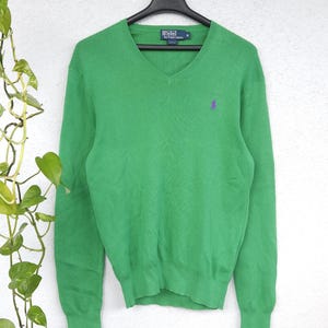 Peut inclure: Pull-over vert vif à col en V et manches longues, suspendu à un cintre noir. Le pull arbore un petit logo violet sur la poitrine. L'étiquette indique "Polo by Ralph Lauren".