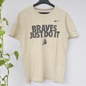 Könnte beinhalten: Beiges Kurzarm-T-Shirt mit dem Text "BRAVES JUST DO IT" in verblichenen schwarzen Buchstaben. Ein kleines Nike-Logo ist über dem Text zu sehen. Das T-Shirt hängt an einem schwarzen Kleiderbügel vor neutralem Hintergrund.