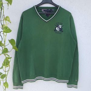 Peut inclure: Pull à manches longues vert avec un col en V et une bordure rayée blanche et marine aux poignets et à l'ourlet. Le pull présente un logo de crête sur la poitrine gauche et une bordure blanche autour de l'encolure. Il est suspendu à un cintre noir.