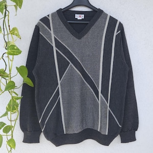 Könnte beinhalten: Ein dunkelgrauer V-Ausschnitt-Pullover mit einem geometrischen Muster in Grau- und Weißtönen. Der Pullover hängt an einem schwarzen Kleiderbügel vor einem weißen Hintergrund. Der Pullover hat lange Ärmel und gerippte Bündchen.