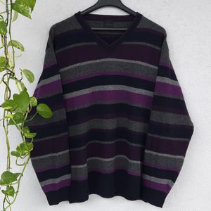 Peut inclure: Un pull à col en V à rayures horizontales violettes, grises et bleu marine. Le pull est suspendu à un cintre noir sur fond blanc. Les manches sont longues et le style général est décontracté.