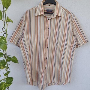 Può includere: Una camicia a maniche corte con abbottonatura frontale e un motivo a righe verticali nei toni del beige, marrone, arancione e bianco. La camicia presenta un colletto classico, una tasca sul petto a sinistra e bottoni scuri. L'etichetta riporta ATTENTION.