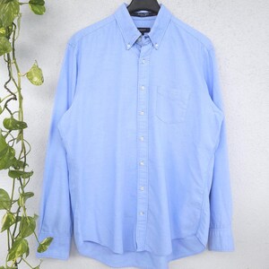 Könnte beinhalten: Ein hellblaues Langarmhemd mit Button-Down-Kragen, einer einzelnen Brusttasche und einer Knopfleiste vorne. Das Hemd hängt an einem schwarzen Kleiderbügel und zeigt den Markennamen Gant auf dem inneren Etikett am Kragen.