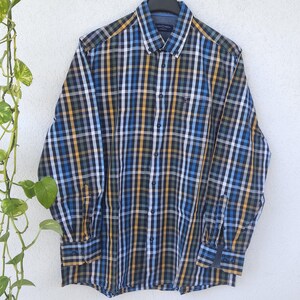 Könnte beinhalten: Ein langärmeliges Hemd mit Knopfleiste und einem Karomuster in Blau, Gelb, Weiß und Dunkelgrau. Das Hemd verfügt über einen klassischen Kragen, eine durchgehende Knopfleiste und geknöpfte Manschetten und hängt an einem schwarzen Kleiderbügel.